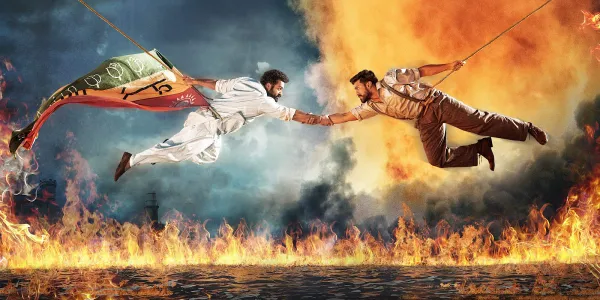 Promo image for RRR featuring two heroes clasping hands while hanging suspended over a fiery river