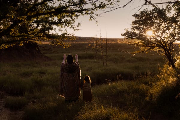 Image from Killers of the Flower Moon. An Osage woman stands with her hands up, palms facing the rays of the setting sun.